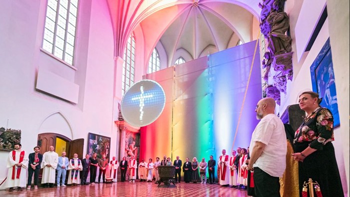 Heller Kirchenraum wird in Regenbogenfarben angestrahlt. Im Kreis stehen Pfarrpersonen u.a.