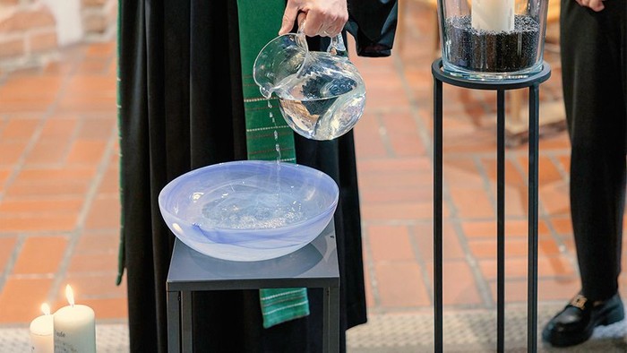 Eine Pfarrperson gießt aus einer Glaskanne wird Wasser in eine hellblaue Taufschale.