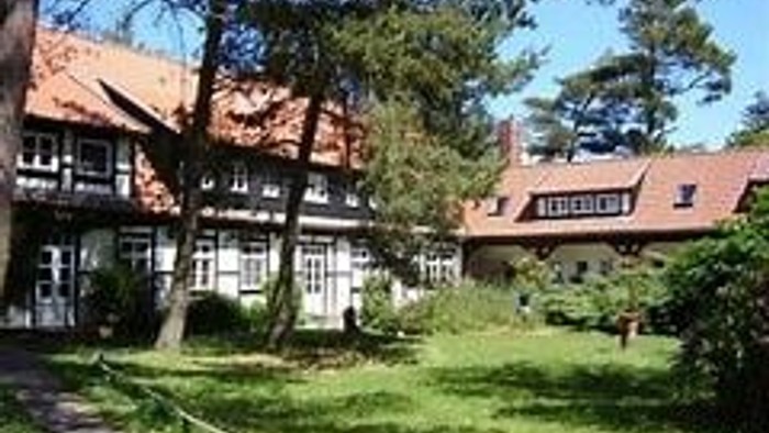 Das Bild zeigt ein großes, traditionelles Haus mit einem roten Dach und weißen Rahmen, umgeben von üppigem Grün und Bäumen.