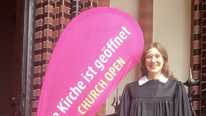 Vikarin Franziska Zellmer steht im Talar vor dem Eingang der Apostel-Paulus-Kirche in Berlin-Schöneberg.