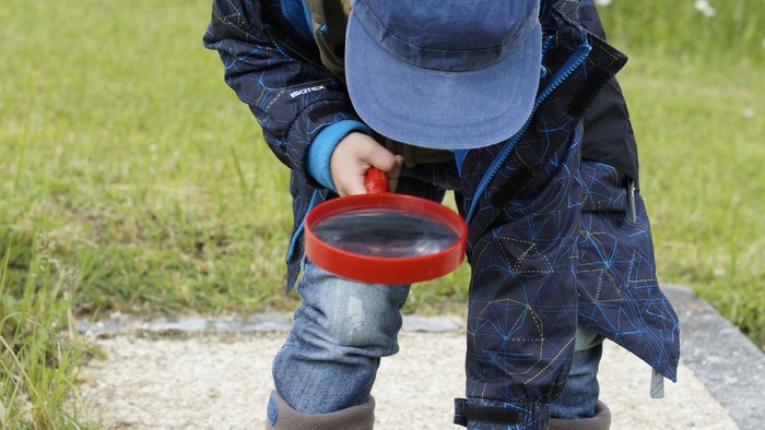 Kleiner Junge sucht auf einem Waldweg mit einer roten Lupe den Boden ab
