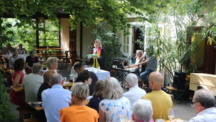 Eine Gruppe von Menschen hört einem Mann mit Gitarre und einer Frau mit Mikrofon zu, die auf einer Bühne unter freiem Himmel spielen.
