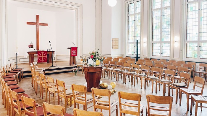 Auf dem Bild ist ein heller Raum zu sehen. Vorne stehen viele Holzstühle im Halbkreis. Hinten ist ein kleiner Altar mit einem großen Holzkreuz. Daneben steht ein Rednerpult mit einem roten Tuch. Der Raum hat große Fenster und viel Licht. In der Mitte steh