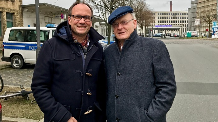Zwei Männer stehen auf einem Stadtplatz, im Hintergrund ein Polizeiwagen.