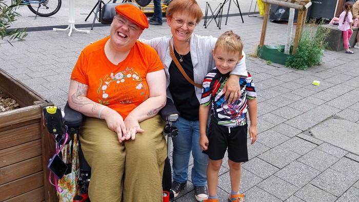 Ein Frau im Rollstuhl mit orangefarbenem Shirt und gleichfarbiger Mütze wird umarmt von einer kleinwüchsigen Frau. Diese hält außerdem einen kleinen Jungen im Arm, ihren Sohn Linus.