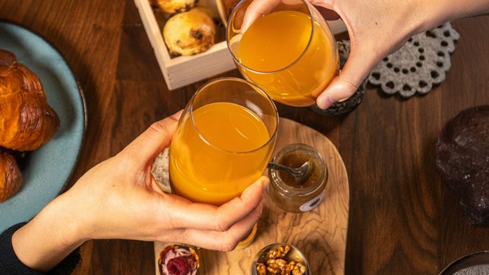 Über einem Tisch, auf dem Gebäck steht, stoßen zwei Hände mit einem Orangensaft an.
