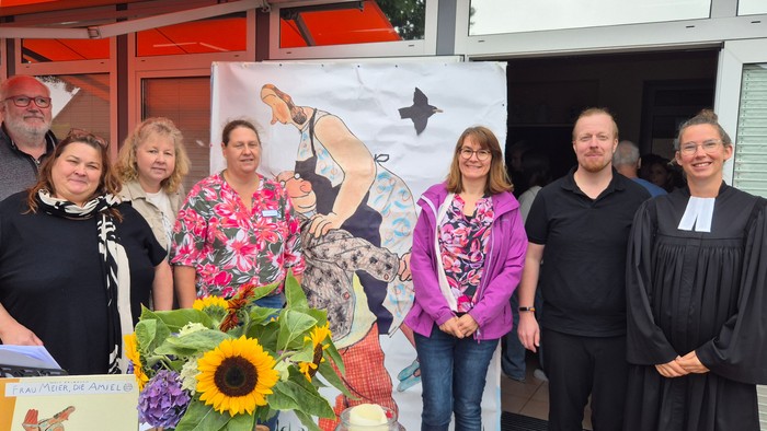 Auf dem Bild sind 7 Menschen aus dem Vorbereitungsteam des Gottesdienstes zu sehen. Sie stehen vor einem Plakat, dass Frau Meier aus der Geschichte 