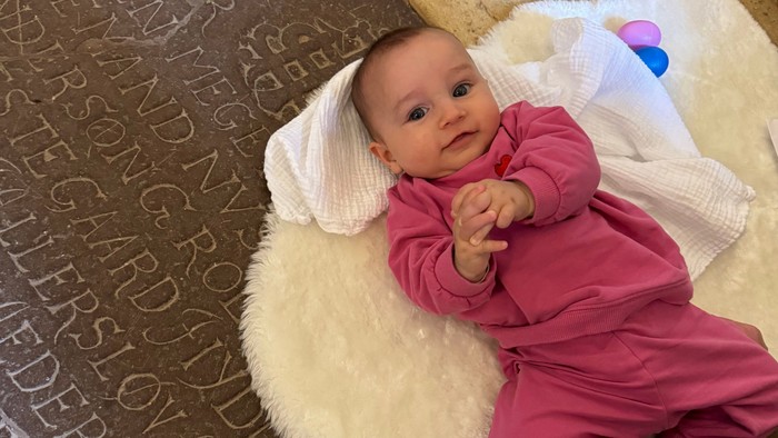 En baby i en lyserød heldragt sidder på en hvid pelset pude og smiler, mens den bliver holdt på et mønstret gulv.