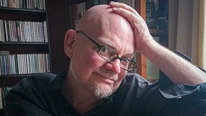 Mann mit Brille sitzt vor Bücherregal und hält den Kopf mit der Hand.