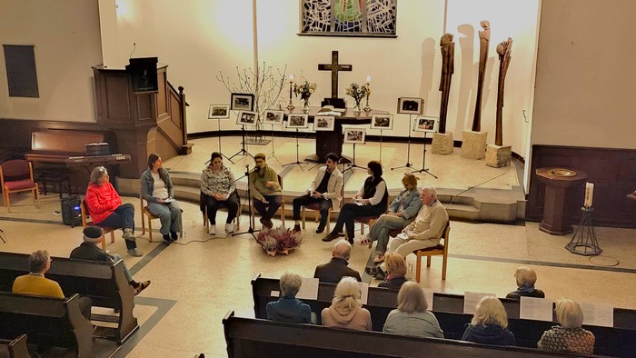 Eine Gruppe von Menschen sitzt in einem Raum, der wie eine Kirche aussieht, und hört einer Person zu, die spricht.