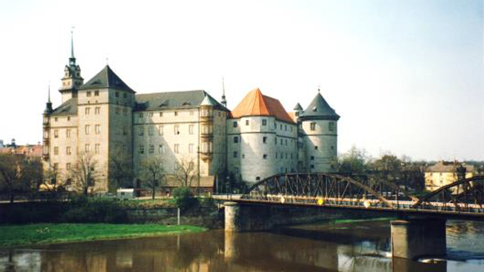 Historische Burg an einem Fluss mit einer Brücke und spiegelnder Wasseroberfläche.