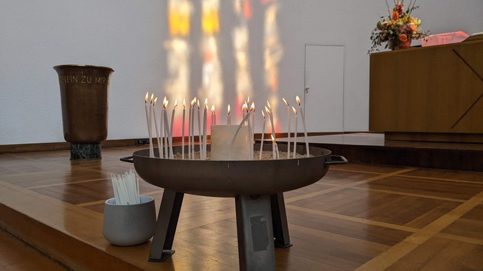 Im Raum stehen mehrere brennende Kerzen in einem Metallbehälter auf einem Tisch.