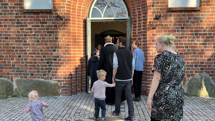 En gruppe af mennesker, heriblandt børn, står udenfor en rød murstensbygning med bogstavelig indgang.