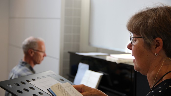 I forgrunden ses en syngende Gitte Friis med en sangbog i hånden. I baggrunden ses lidt sløret organist Lars Christesen, der spiller på klaveret.