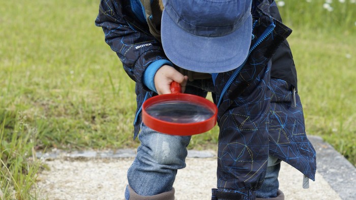 Kleiner Junge sucht auf einem Waldweg mit einer roten Lupe den Boden ab