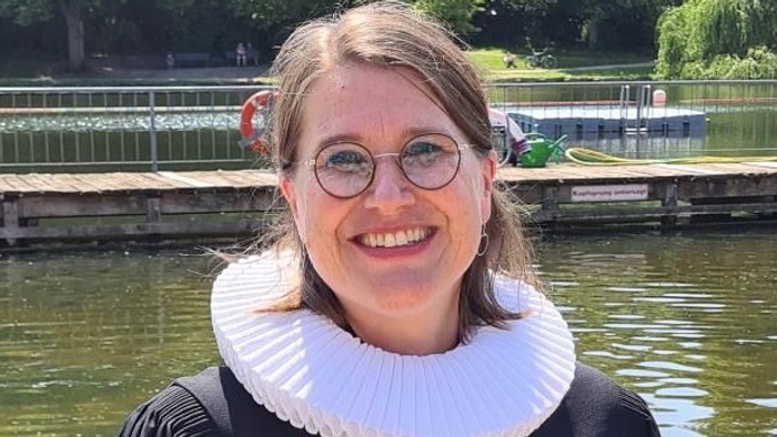 Eine Frau in Abschlusskleidung steht am Flussufer und lächelt.