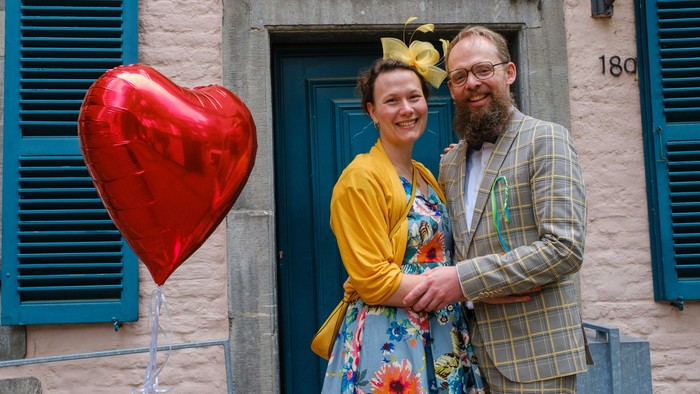 Ein Paar steht vor einem blauen Tür, die Frau hält einen roten Herzballon.