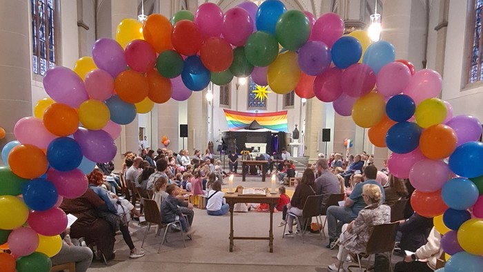 Farbenfrohe Ballonbogen in Kirche, Menschen sitzen auf Stühlen, Musikgruppe spielt