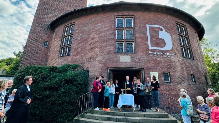 Die Lukaskirche in Eckesey fällt besonders durch ihre ovale Form auf. Menschen stehen vor dem Eingang der Kirche und links und rechts der Treppe, um in einem Entwidmungsgottesdienst Abschied zu nehmen.