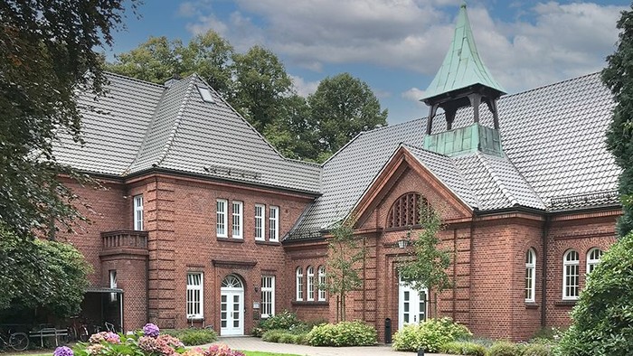 Ein rotes Backsteingebäude mit spitzen Dächern und einem grünen Turm, umgeben von einem gepflegten Garten mit blühenden Blumen.