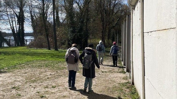 4 Menschen mit Rucksäcken laufen neben einer hohen Mauer entlang. Im Hintergrund Bäume und ein Fluss.