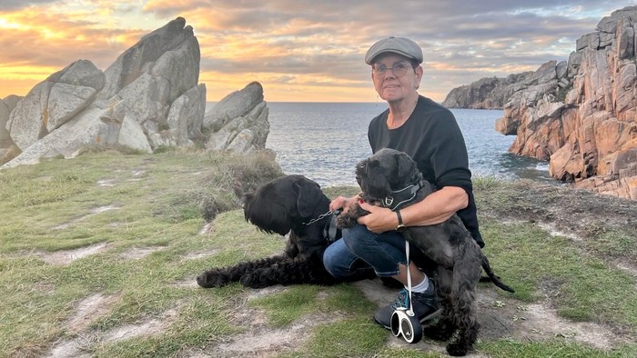 Ein Mann kniet mit zwei Hunden an einem felsigen Strand bei Sonnenuntergang