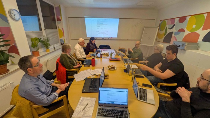Eine Gruppe von Menschen sitzt um einen Tisch mit Laptops in einem Klassenzimmer.