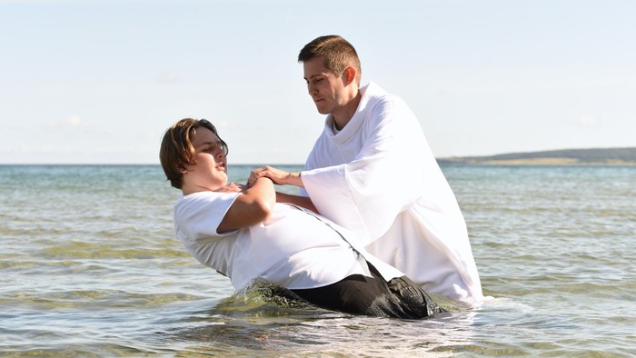 Ein Mann in weißem Gewand tauft einen Jungen im Wasser.