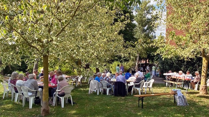 Mehrere Tischgruppen mit älteren Menschen unter Bäumen 