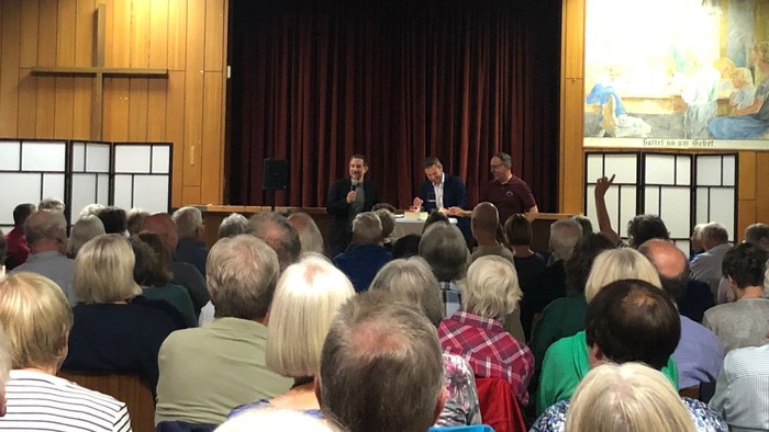 Eine Gruppe von Menschen hört einem Redner auf einer Bühne zu.