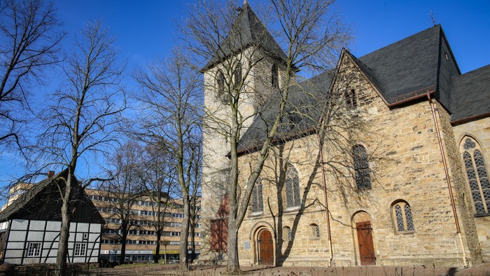 Alte Kirche mit hohem Turm und spitzen Fenstern, umgeben von kahlen Bäumen und modernen Gebäuden im Hintergrund