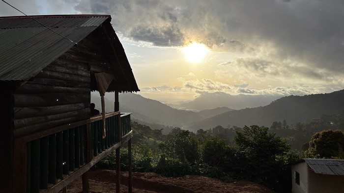 Ein Panoramablick über die Usambaraberge im Sonnenuntergang. Links ein Holzhaus mit Veranda.