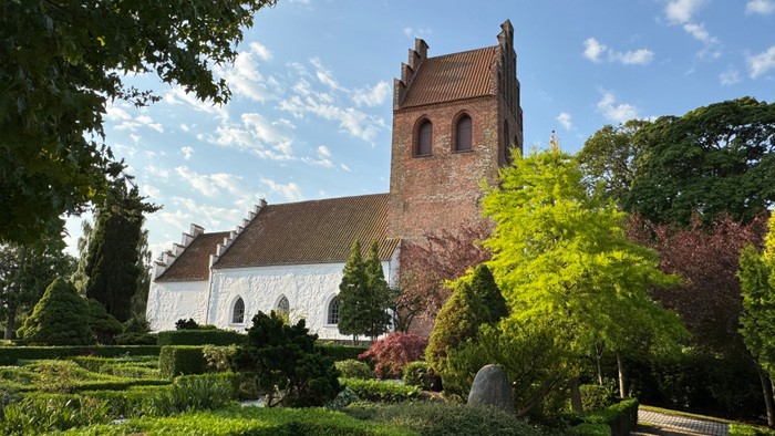 En gammel kirke med tårn omgivet af grønne træer og buske