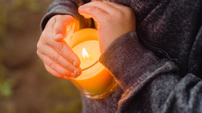 Eine brennende Kerze in einem gelbschimmernden Glas wird von zwei Händen gleichsam getragen und vor Wind geschützt. Ein Gesicht ist auf dem Bild nicht zu sehen.