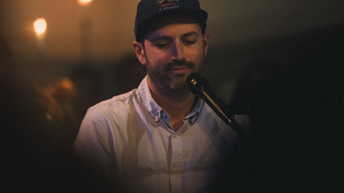 Musikeren S. Carey står ved en mikrofon og spiller i dæmpet belysning under en intim koncert. Han har en hvid skjorte og mørk kasket på. Slørede publikumsfigurer og stearinlys ses i baggrunden. Foto: Sam Leitch