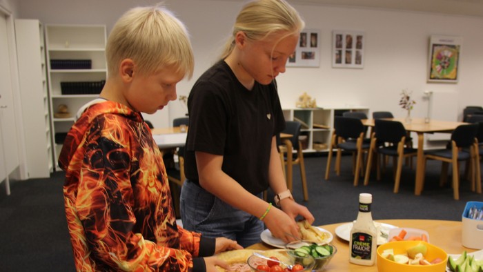 To børn står ved et bord , hvor der står skåle med frugt og grøntsager. De har hver deres pandekage, som de lægger fyld på.