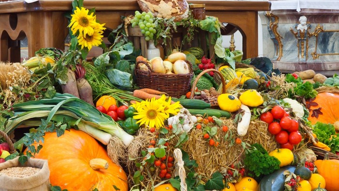Ein großer Korb gefüllt mit einer Vielzahl von frischem Obst und Gemüse, darunter Kürbisse, Äpfel, Tomaten und Blumen, steht auf einem Tisch.