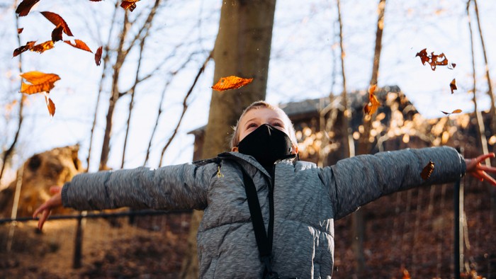 Ein Kind mit bedecktem Mund und grauer Jacke steht mit ausgestreckten Armen im Freien, während um es herum Herbstblätter fallen.