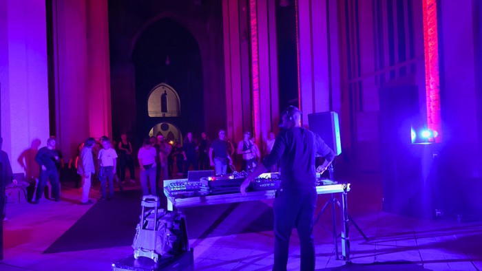 Bunt angestrahlter Kirchenraum, Menschen tanzen, im Vordergrund ein DJ-Pult.