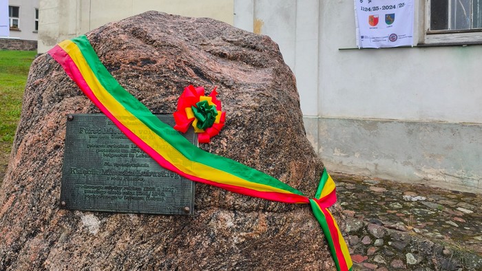 Großer Findling mit Gedenkplatte umwunden von grün-gelb-rotem Band vor einer Kirche.