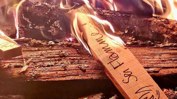 Flamme eines Lagerfeuers, darin ein Holzschild mit Aufschrift