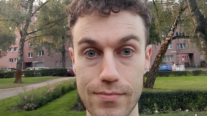 Eine Person steht lächelnd draußen in einem Park mit Bäumen und Gebäuden im Hintergrund.