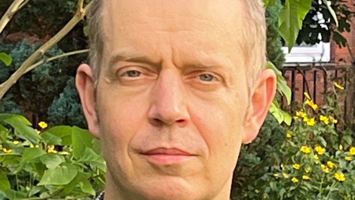 Ein Mann mit kurzem Haar trägt ein kariertes Hemd und einen braunen Blazer im Freien mit Grünflächen im Hintergrund.