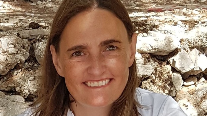 Eine lächelnde Frau in einem weißen Hemd posiert im Freien vor einem steinernen Mauerhintergrund.