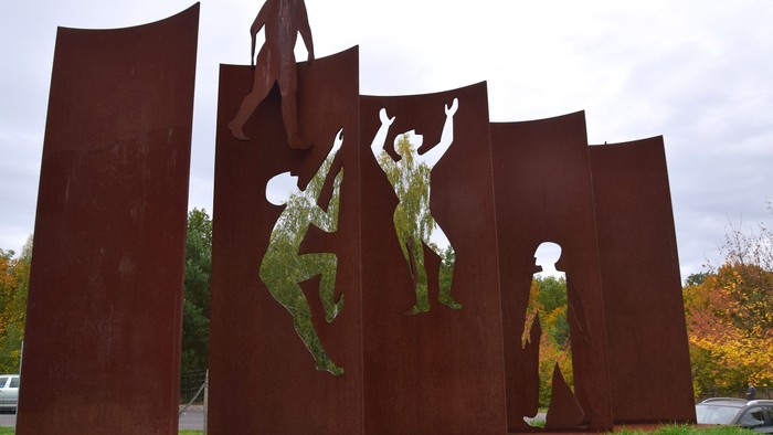 Die Skulptur zeigt mehrere Figuren in dynamischen Posen auf rostfarbenen Metallplatten.
