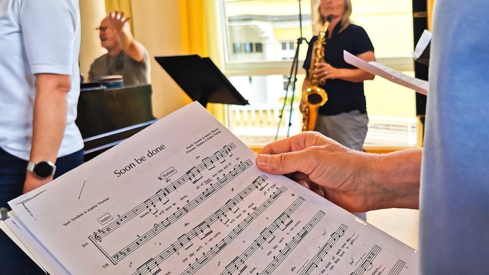Nahaufnahme eines Notenblatts bei der Probe mit einer Saxophonspielerin und dem Chorleiter im Hintergrund