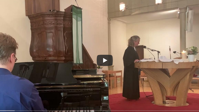 Ein Mann steht hinter einem Pult und spricht in einen Mikrofon, während ein anderer Mann ein Klavier spielt.