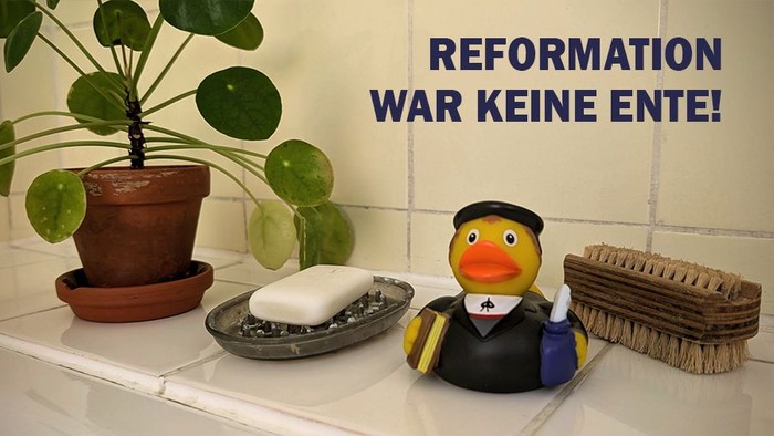 Luther-Gummiente mit Bibel und Feder neben Seife und Bürste am Badewannenrand. Spruch 