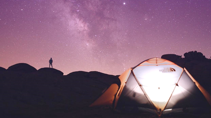 Einzelne Person steht unter sternenklarem Himmel neben einem beleuchteten Zelt