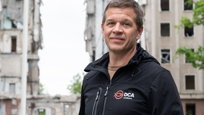 Mand i sort jakke med DCA logo står foran skadede bygninger